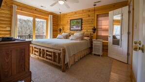 A log cabin bedroom with a bed, a ceiling fan, a painting, and a view of the outdoors.