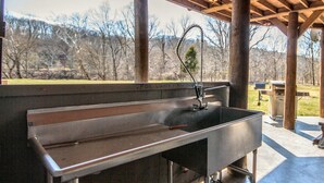 A stainless steel outdoor kitchen with a built-in sink and faucet, located under a wooden pergola with a view of a wooded area.