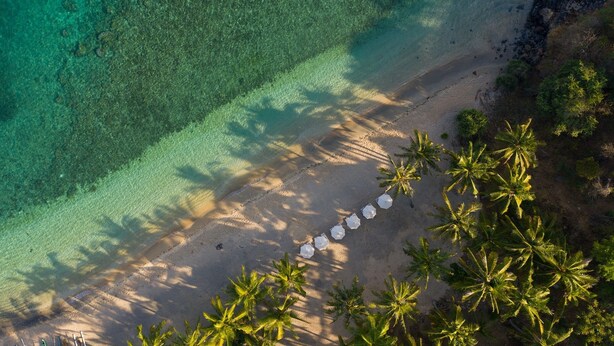 An aerial view of a beach with clear turquoise water, a sandy shore, and a row of umbrellas.