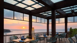 A covered outdoor seating area with tables and chairs, overlooking a sunset over the sea.
