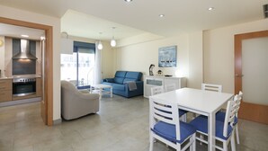 Eine moderne Küche mit eingebauten Geräten, ein Essbereich mit weißem Tisch und blauen Stühlen, ein blaues Sofa und ein Bild von Segelbooten an der Wand.