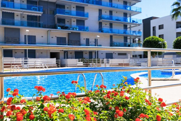 A swimming pool with a metal railing, surrounded by red flowers, and a multi-story building with balconies in the background.