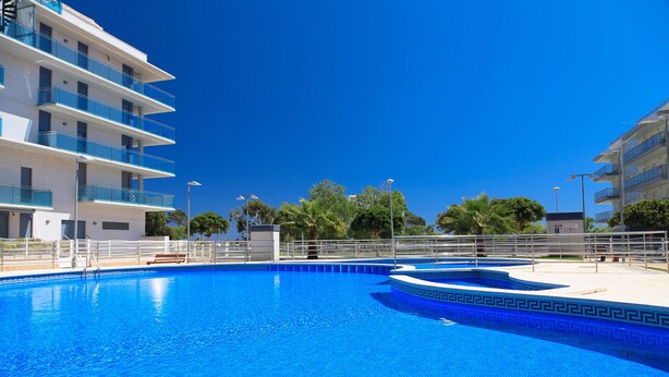 Ein Schwimmbecken, umgeben von modernen Gebäuden mit Balkonen, blauem Himmel und etwas Grün.