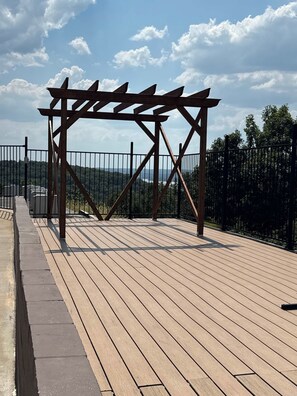 Una terraza de madera con estructura de pérgola, rodeada por una valla metálica, con vistas a un paisaje montañoso.