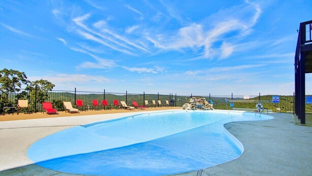 A swimming pool with a curved edge, surrounded by a fence. There are lounge chairs along the poolside. - 24-6 Serenity Escape Penthouse w/Indoor/Outdoor Pool / Splash Pad Near SDC (Branson)