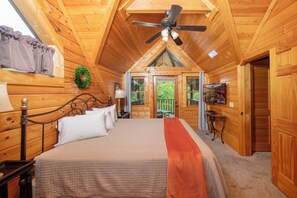 A wooden cabin bedroom with a bed, ceiling fan, and a view of the outdoors.