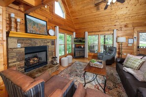 A log cabin interior with a stone fireplace, leather furniture, and large windows.