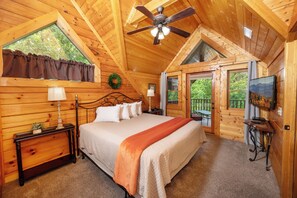 A wooden cabin bedroom with a bed, nightstands, a ceiling fan, a TV, and a window with curtains.