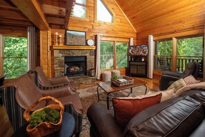 L’intérieur d’une cabane en rondins, avec une cheminée en pierre, des canapés en cuir et de grandes fenêtres donnant sur la forêt.
