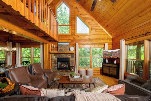 A spacious living room with a fireplace, large windows, and a high ceiling.