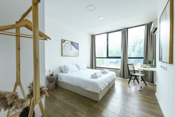 A modern bedroom with a large bed, a wooden hanger, a desk with a chair, and a window with curtains.