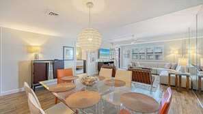 A modern living room with a glass dining table, orange chairs, a sofa, and a large chandelier. - Spectacular Beach View Steps from Siesta Key Beach! (Sarasota)