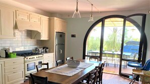 Une cuisine avec des meubles blancs, une cuisinière et un réfrigérateur. On y trouve une table à manger avec des chaises et une grande fenêtre surmontée d’un arc.