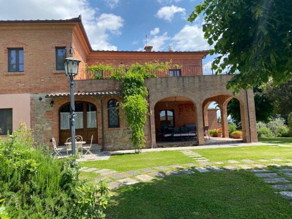 Un bâtiment de deux étages en briques, doté d’une cour en pierre, d’un espace extérieur aménagé avec des sièges et d’une pergola couverte de plantes grimpantes.