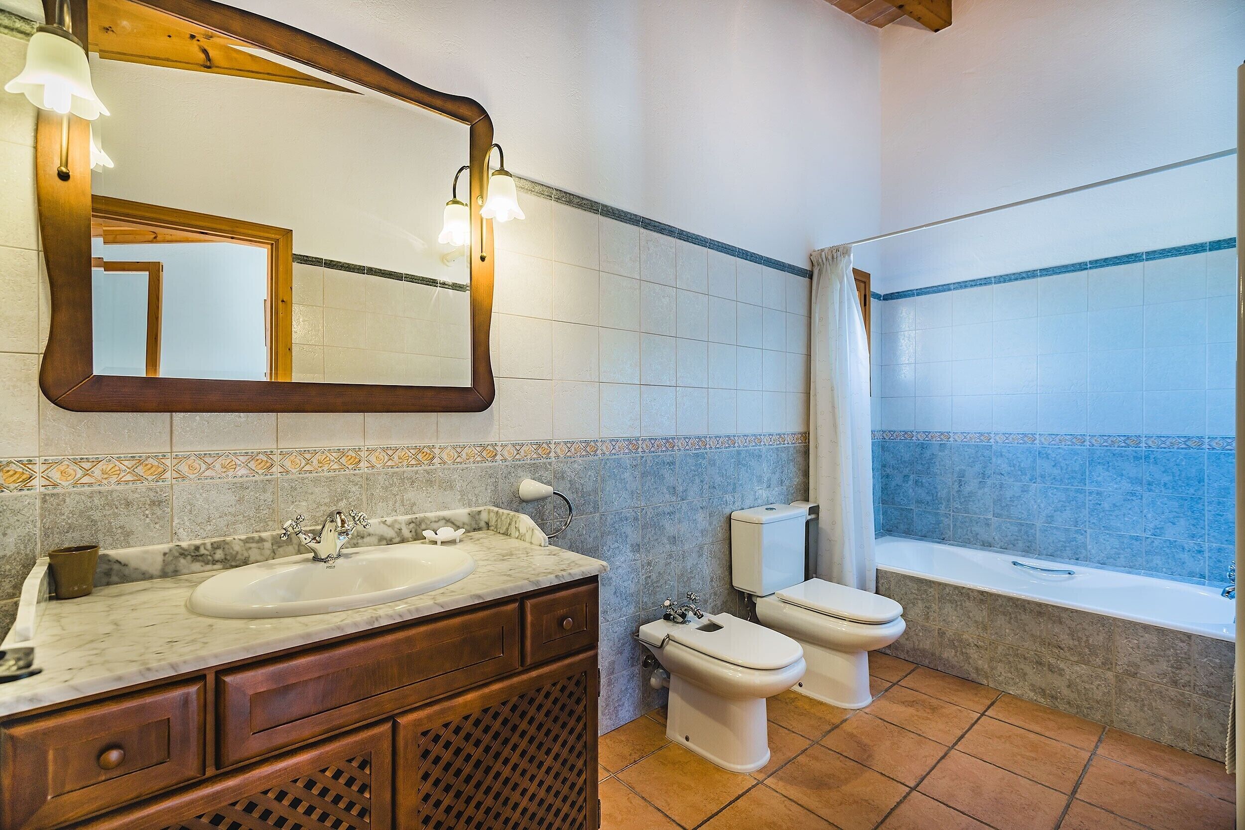 A bathroom with a marble sink, wooden vanity, two toilets, a bathtub, and a large mirror with lights.