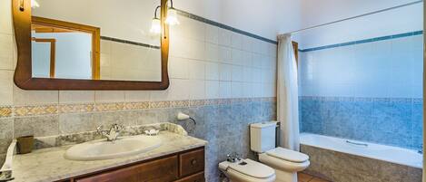 A bathroom with a marble sink, wooden vanity, two toilets, a bathtub, and a large mirror with lights.