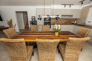 Un comedor con una amplia mesa de madera rodeada de sillas tejidas, una cocina al fondo y una moderna lámpara colgante.
