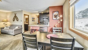 Un salón acogedor con un sofá gris, una mesa de comedor de madera con cuatro sillas, una cocina con armarios oscuros y un microondas, y un ventanal con vistas a un paisaje nevado.