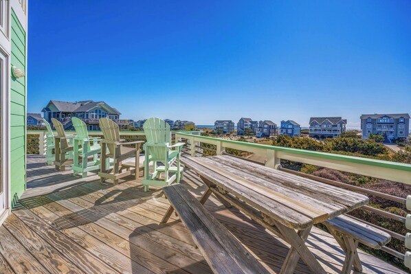 A wooden deck with green Adirondack chairs and picnic tables overlooking a beachfront community with colorful houses.