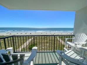 A balcony with white chairs overlooking a beach with people and umbrellas.
