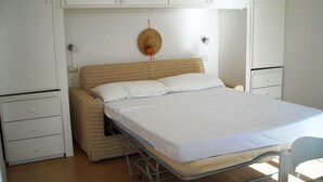 A single-bed room with a metal frame, a beige mattress, and a white sheet. There is a small white cabinet to the left and a white wardrobe to the right. - Cozy Studio Erato ideal for a couple (Menfi)