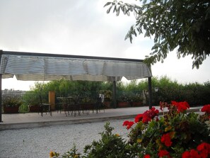 Ein überdachter Außenbereich mit Metallstühlen und -tischen, umgeben von Pflanzen und Blumen.