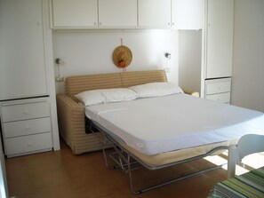 Ein Zimmer mit einem Einzelbett, einem Metallgestell, einer beigen Matratze und einem weißen Bettlaken. Links befindet sich ein kleiner weißer Schrank und rechts ein weißer Kleiderschrank.