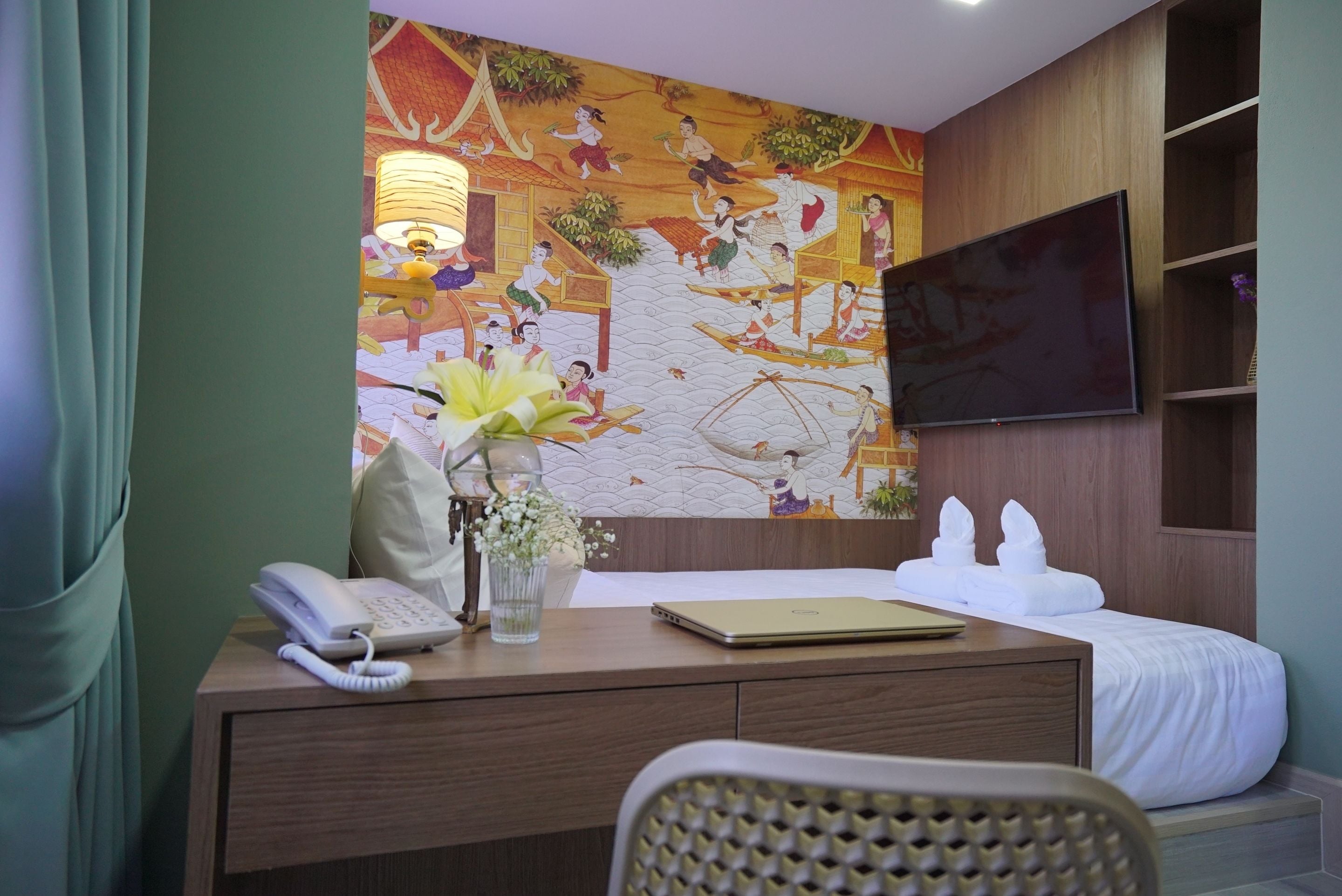 A hotel room with a bed, a desk with a phone and a laptop, a TV mounted on the wall, and a mural depicting traditional scenes.