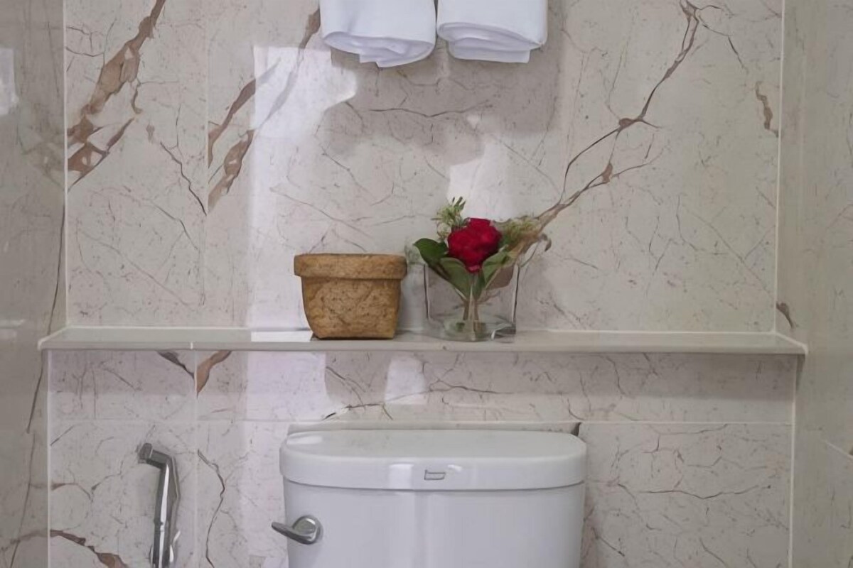 A bathroom with a marble wall, a toilet, a shelf with a flower vase, and a shower area.