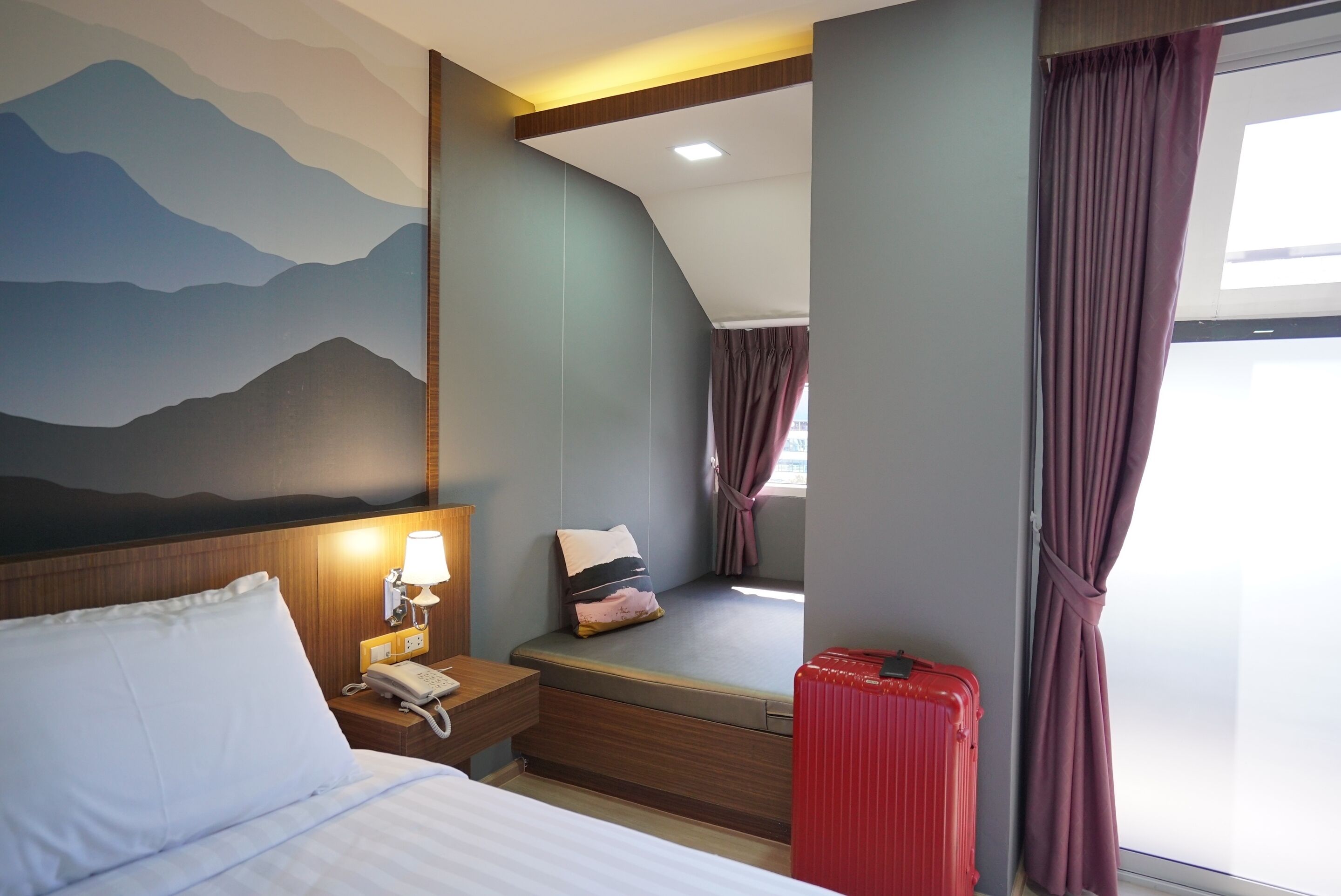 A hotel room with a bed, bedside table with a lamp, a window with curtains, a red suitcase, and a wall mural.