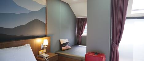 A hotel room with a bed, bedside table with a lamp, a window with curtains, a red suitcase, and a wall mural.
