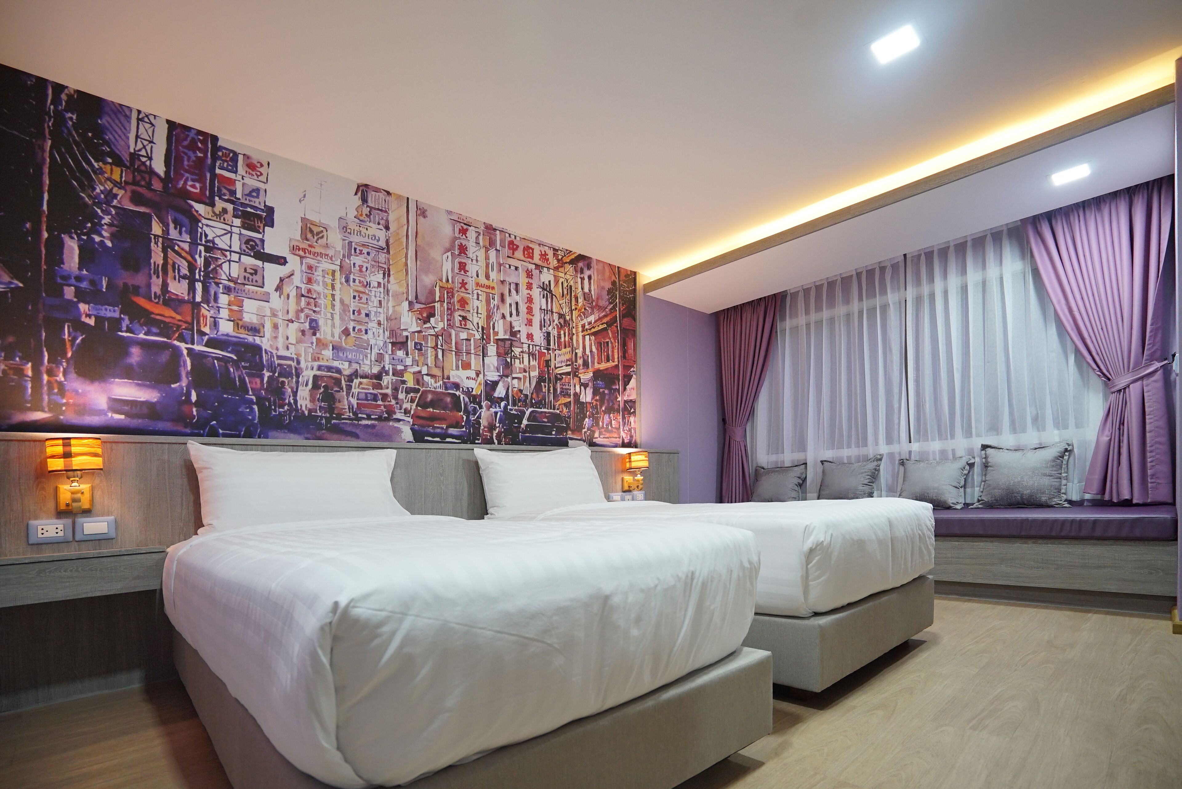 Quarto de hotel com duas camas, um mural grande com vista para a cidade na parede, uma janela com cortinas roxas e um banco embutido com almofadas.