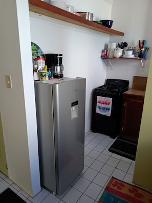 A compact kitchen with a stainless steel refrigerator, a gas stove, and a microwave. There are shelves above the stove holding various kitchen items.