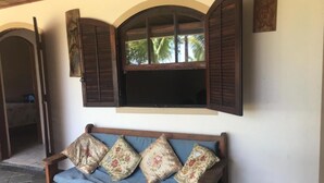 Un banco de madera con cojines florales, un televisor montado en una pared con contraventanas de madera y un suelo de piedra.