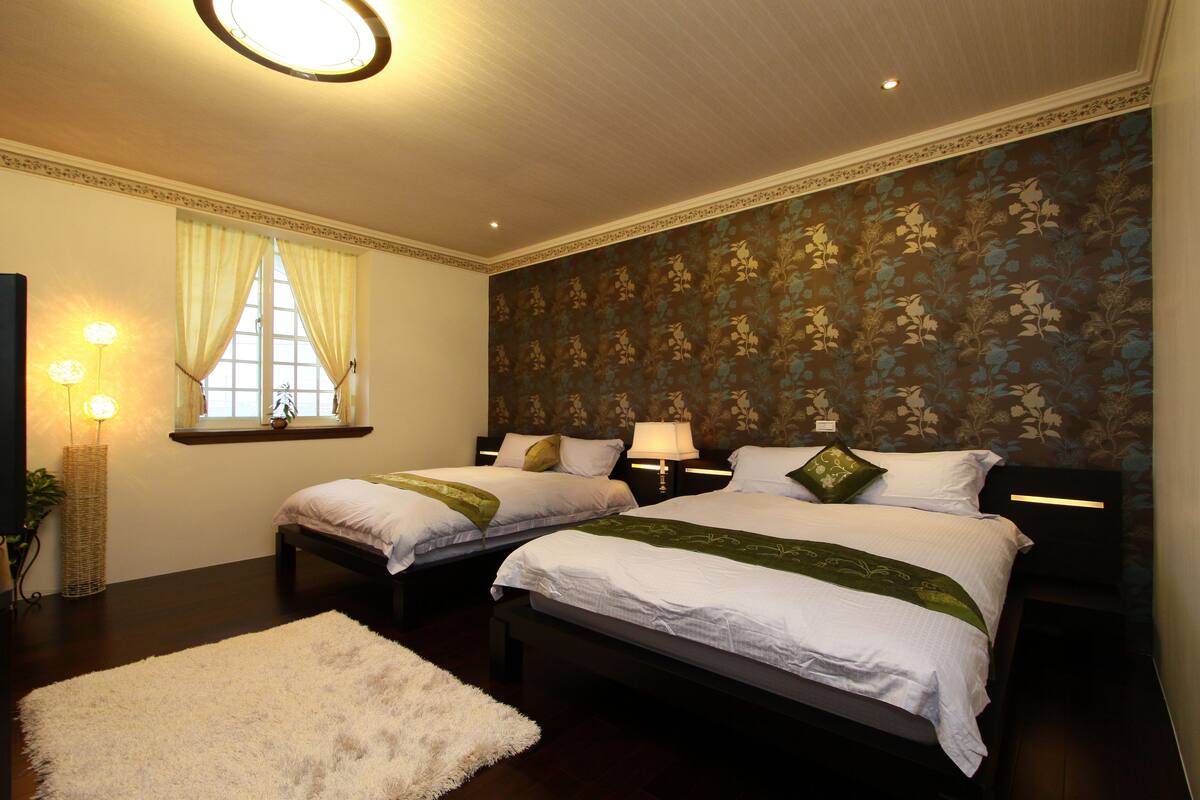Habitación de hotel con dos camas, un ventanal con cortinas, una pared con estampado floral y una lámpara de pie.