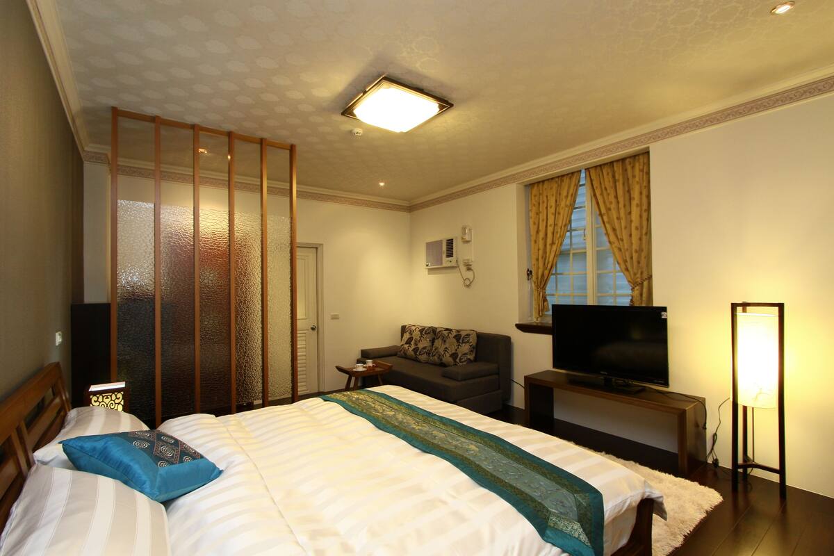 Una habitación de hotel con una cama, televisión, lámpara, sofá y ventana con cortinas.