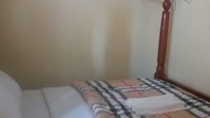 A single bed with a checkered bedspread and a white pillow. A wooden nightstand with a lamp is beside the bed. - Kabz Hotel (Kampala)