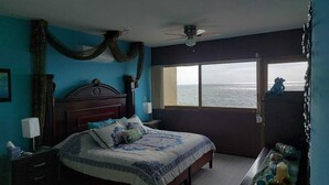 A bedroom with a bed, a nightstand, a dresser, a window with a view of the ocean, and a ceiling fan. - Beautiful Private Beachfront Villa (Nuevo Mazatlan)