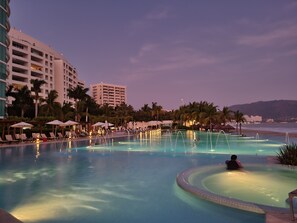 Una amplia piscina al aire libre con sillas de descanso y sombrillas, rodeada de palmeras y edificios de hotel.