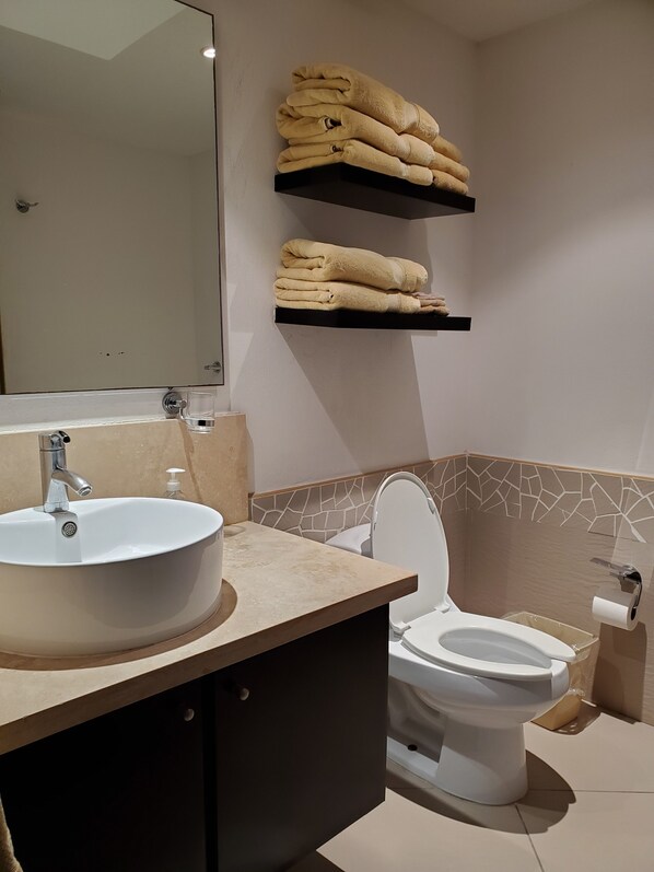 Un baño con un lavabo blanco, un inodoro y un espejo. Hay dos repisas con toallas dobladas sobre el lavabo.