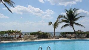 A clear blue pool with a metal ladder, surrounded by lounge chairs and palm trees, with the ocean in the background. - Magnificent Beach front condo with pool and restaurant/bar (Freeport)