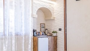 Un couloir étroit avec un mur en briques blanches, une porte en bois et un petit coin cuisine comprenant un réfrigérateur, une machine à café et une plante en pot.