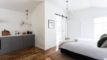 Relax in this serene bedroom with a large bed against a white wall, adjacent to a compact kitchen area with grey cabinetry.