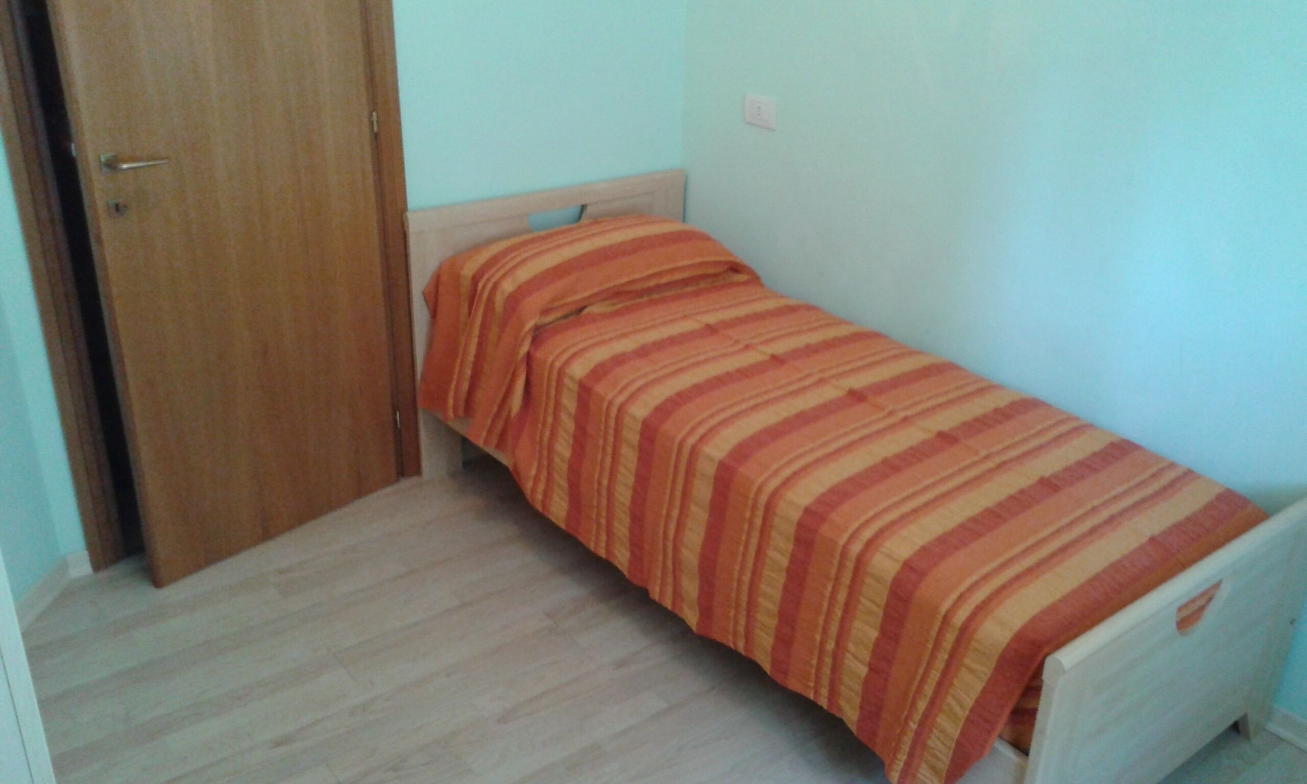 A small, single-bed room with a wooden door and a plain light-colored wall.