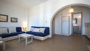 Ein modernes Wohnzimmer mit einem blauen Sofa, weißen Sessel und einem Couchtisch. Ein Bogen führt in den Küchenbereich.