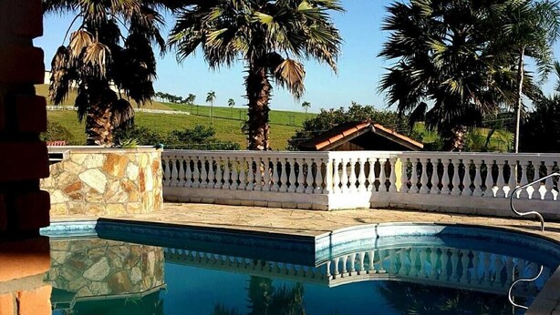 A swimming pool with a stone border and a white balustrade, surrounded by palm trees and a view of a grassy area. - Beautiful house with the best View (Sao Roque)