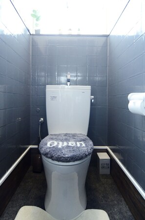A small, dark-colored bathroom with a toilet, a trash bin, and a small box labeled "KEEP YOUR HOME CLEAN." - South Island (Itoman)