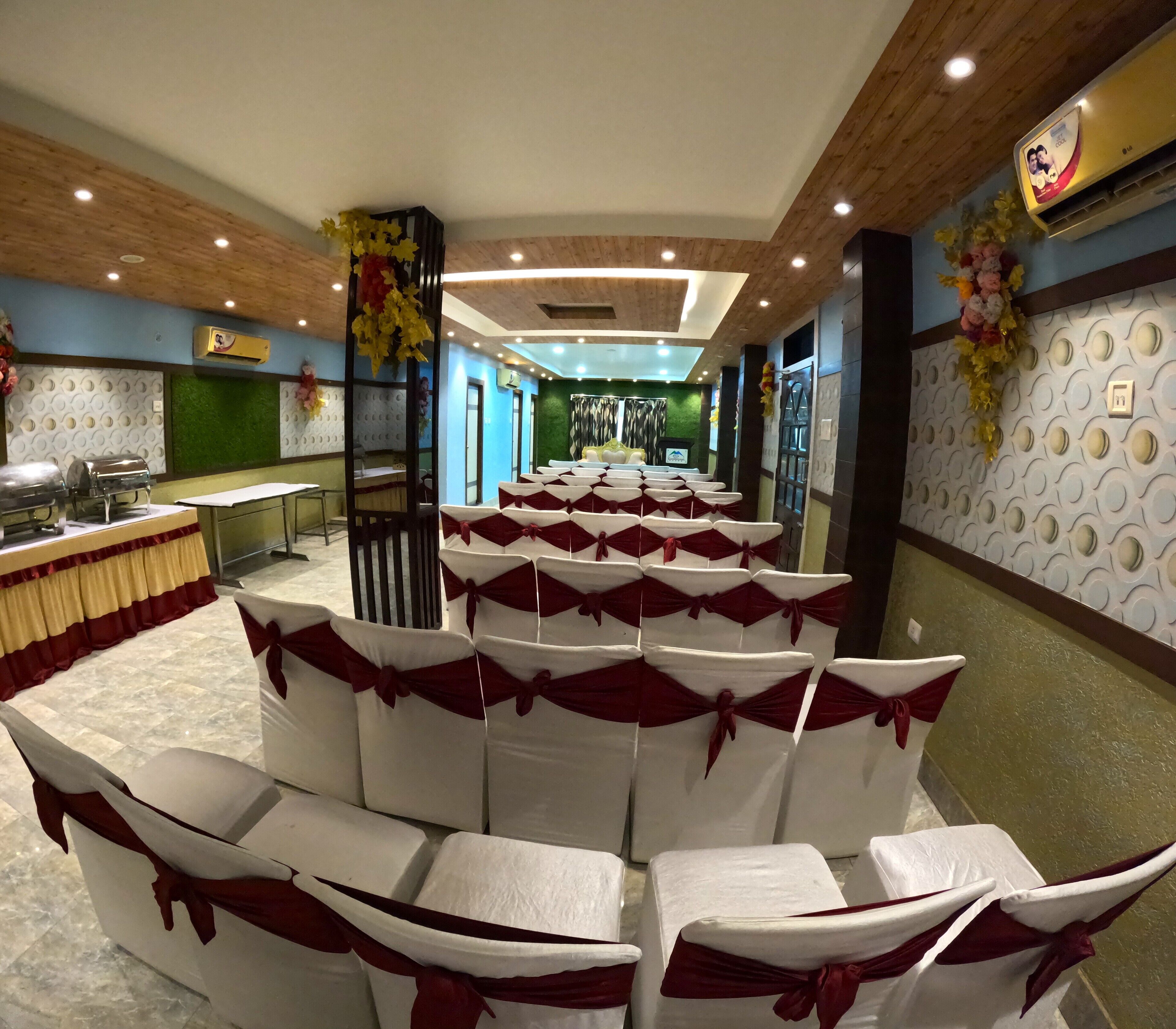 A banquet hall with rows of white chairs with red sashes, a buffet area, and a stage with a green backdrop.