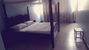 Una cama con dosel, con una colcha y almohadas blancas, un taburete de madera pequeño y un suelo de baldosas.