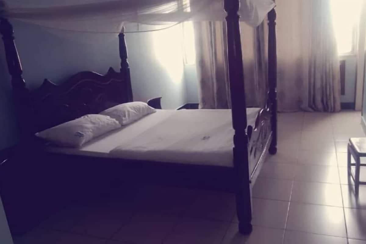 A four-poster bed with a white bedspread and pillows, a small wooden stool, and a tiled floor.